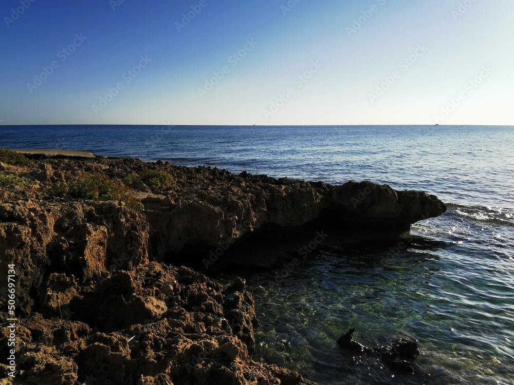 Long-hardened lava, porous, sharp coast of the Mediterranean Sea with incoming waves against a blue cloudless sky. and sea water.