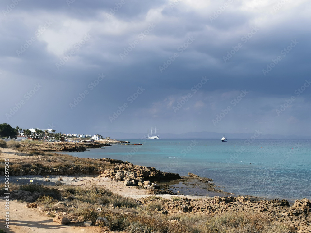 Sunny coast of the Mediterranean Sea with  stones, dramatic sky and rain above the sea, boats in the sea and the largest sailing yacht in the world.