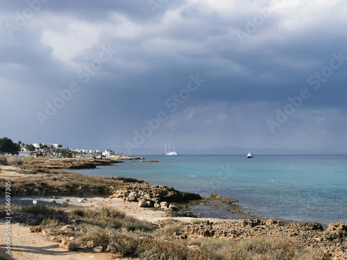 Sunny coast of the Mediterranean Sea with  stones, dramatic sky and rain above the sea, boats in the sea and the largest sailing yacht in the world. photo