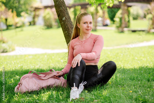 Pretty slim fit blond woman in sportswear and sportsbag sitting and resting on the lawn in the park photo