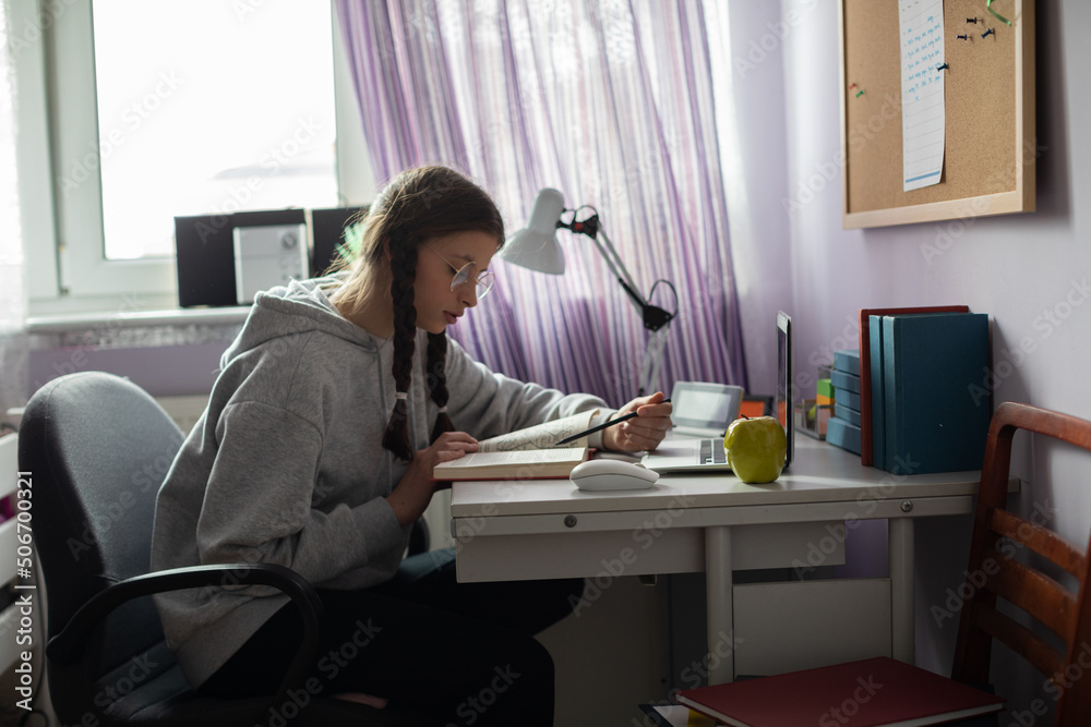 A teenage girl with glasses reads a book at her desk at home. A teenage girl with glasses reads a book at her desk at home.