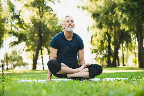 Caucasian mature male yogi sitting in lotus position on fitness mat on green grass in local park forest outdoors. Healthy lifestyle.