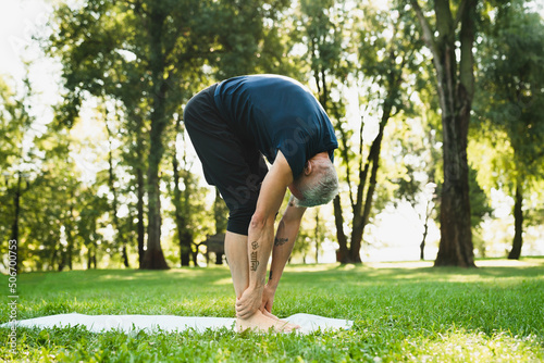 Caucasian mature man bending in sporty clothes on fitness mat in public park outdoors. Active seniors, healthy lifestyle. Stretching concept