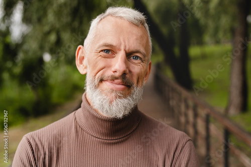 Closeup cropped portrait of a smiling confident caucasian handsome mature man looking at the camera while walking outdoors in city park forest.