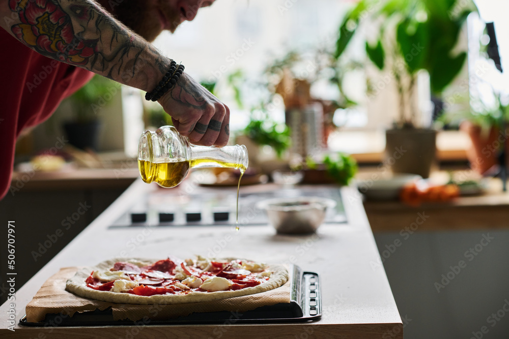 Young bearded man cooking pizza in kitchen at home adding some olive oil over it before putting it into oven