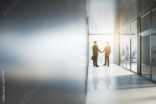 Business partnership concept with man handshaking in sunny office hall with city view and blank blurry wall with copyspace. Mock up, close up