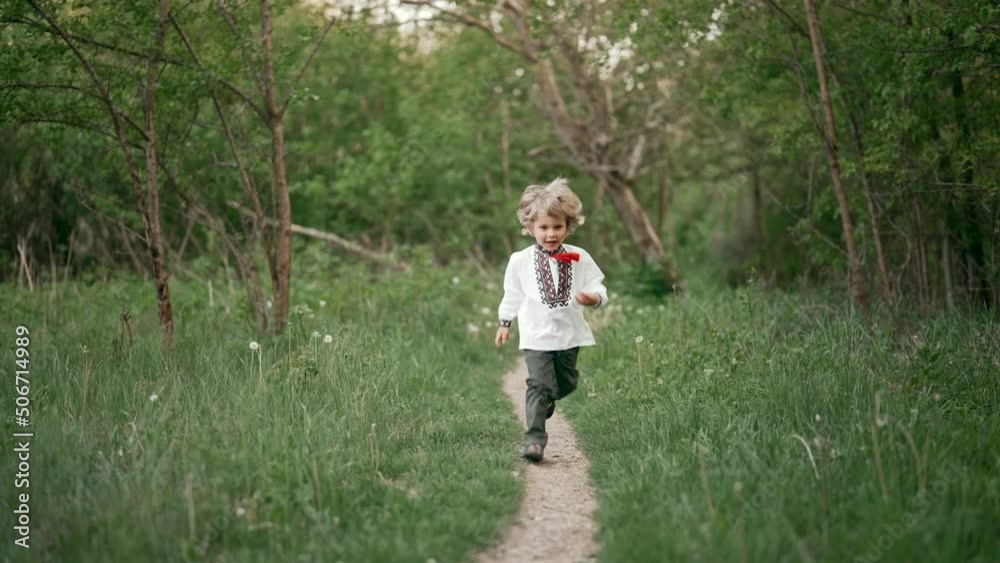 Little ukrainian boy cheerfully running along path on forest lawn or in park. Child in traditional embroidery vyshyvanka shirts. Ukraine, freedom, national costume, patriot Little ukrainian boy cheerfully running along path on forest lawn or in park. Child in traditional embroidery vyshyvanka shirts. Ukraine, freedom, national costume, patriot