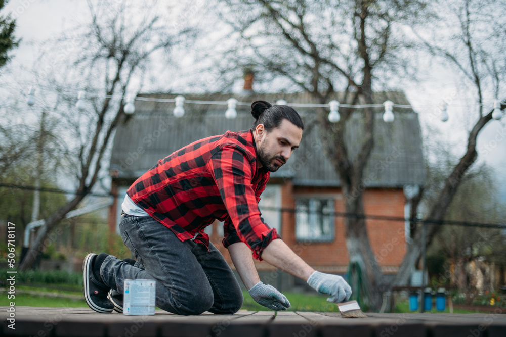A young man in home clothes paints a wooden deck in the garden with a brush. A young man in home clothes paints a wooden deck in the garden with a brush.
