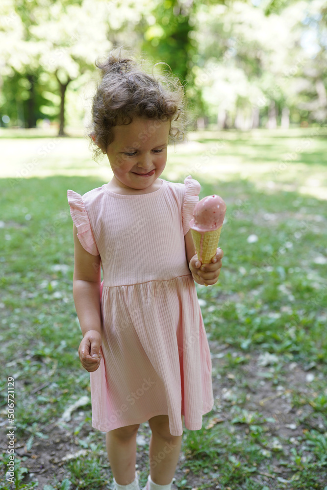 Cute little girl child in pink dress standing on green grass and eating icecream Cute little girl child in pink dress standing on green grass and eating icecream