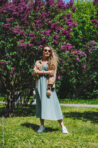 A beautiful curly woman in a summer dress runs and rejoices, she is wearing sunglasses. Against the background of a purple lilac bush