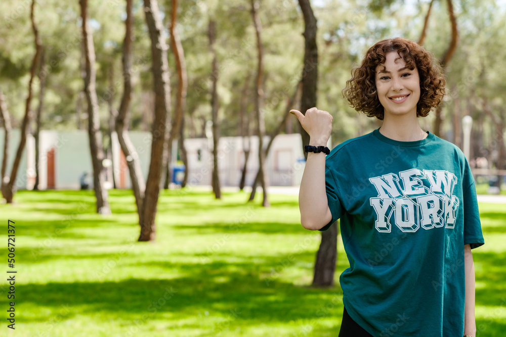 Young redhead woman wearing green tee standing on city park, outdoors smiling with happy face looking at the camera and pointing to the side with thumb up. Showing advertisement, indicating aside.
