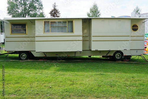 Großer weißer Wohnwagen mit goldenen Streifen auf  grüner Wiese vor Bäumen und Himmel bei Sonne im Frühling 