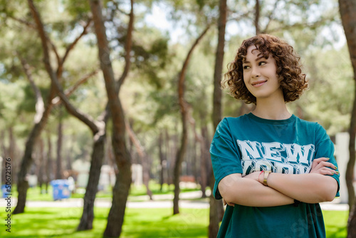 Young redhead woman wearing green t-shirt standing on city park, outdoors. Happy face smiling with crossed arms looking away with confident expression. Positive person, feels satisfied. Copy space.