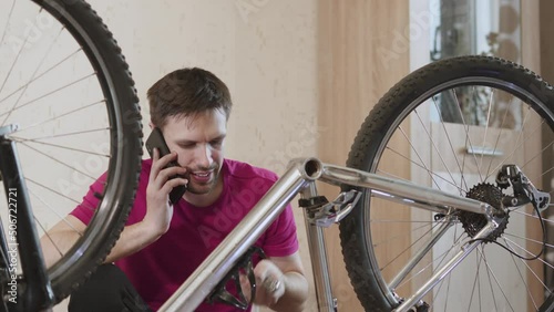 Man sits near dismantled inverted bicycle in room and calls the service. Man puts metal rod through hole in frame and picks up chainrings. Frustrated guy shrugs his shoulders in incomprehension. photo