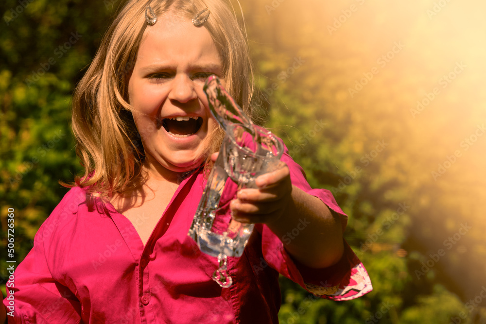 Fototapeta premium The child splashes water from a glass in a rage . A young girl screams and swears. Child aggression .