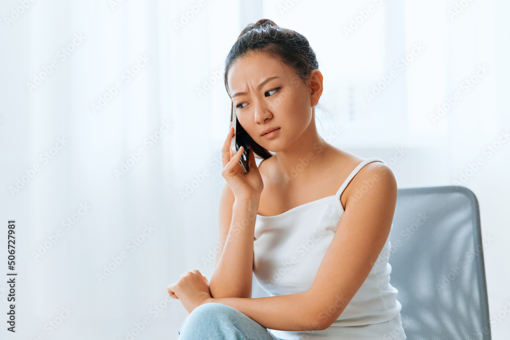 Wary irritated angry tanned lovely young Asian woman listen bad news at call using phone at home interior living room. Distance communication Social media concept. Cool offer Banner Wide angle Wary irritated angry tanned lovely young Asian woman listen bad news at call using phone at home interior living room. Distance communication Social media concept. Cool offer Banner Wide angle