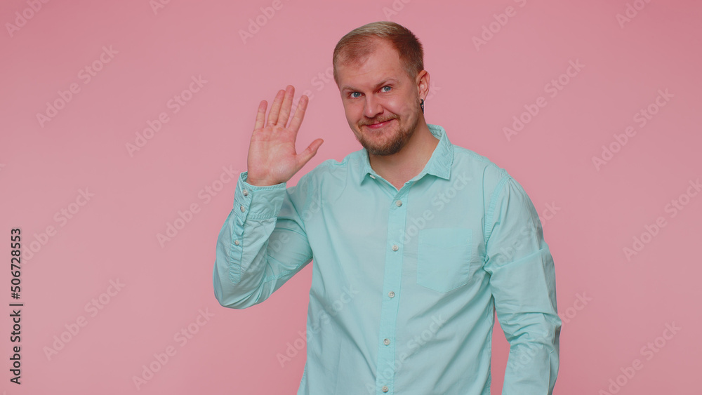 Handsome bearded man in shirt smiling friendly at camera and waving hands gesturing hello or goodbye, welcoming with hospitable expression. Young happy guy indoors isolated on pink wall background Handsome bearded man in shirt smiling friendly at camera and waving hands gesturing hello or goodbye, welcoming with hospitable expression. Young happy guy indoors isolated on pink wall background