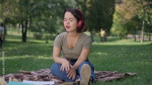 Joyful little woman in headphones listening to music dancing in slow motion sitting on balnket in park outdoors. Wide shot portrait of Caucasian person with dwarfism enjoying hobby on weekend photo