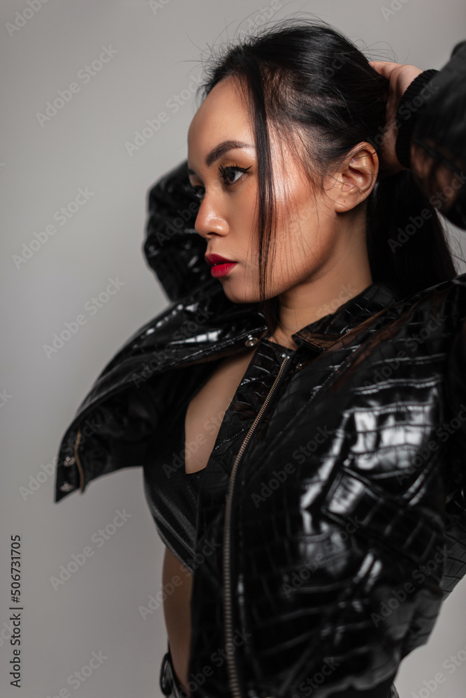 Fashionable beautiful sexy Asian woman in black stylish leather clothes with jacket and sconces in the studio fixing her hair. Glamour pretty girl Fashionable beautiful sexy Asian woman in black stylish leather clothes with jacket and sconces in the studio fixing her hair. Glamour pretty girl