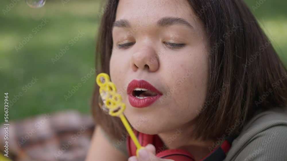 Close-up cheerful young woman with dwarfism blowing soap bubbles laughing looking at camera. Headshot of Caucasian little person posing in slow motion enjoying leisure in park outdoors Close-up cheerful young woman with dwarfism blowing soap bubbles laughing looking at camera. Headshot of Caucasian little person posing in slow motion enjoying leisure in park outdoors