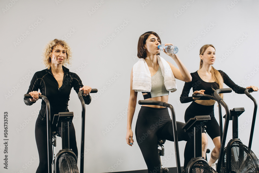 Young women drinking water and using air bike for cardio workout at cross training gym