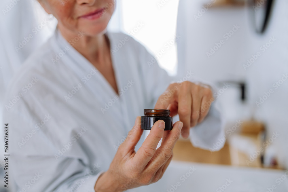 Senior woman in bathrobe applying natural face cream in bathroom, skin care and morning routine concept. Senior woman in bathrobe applying natural face cream in bathroom, skin care and morning routine concept.