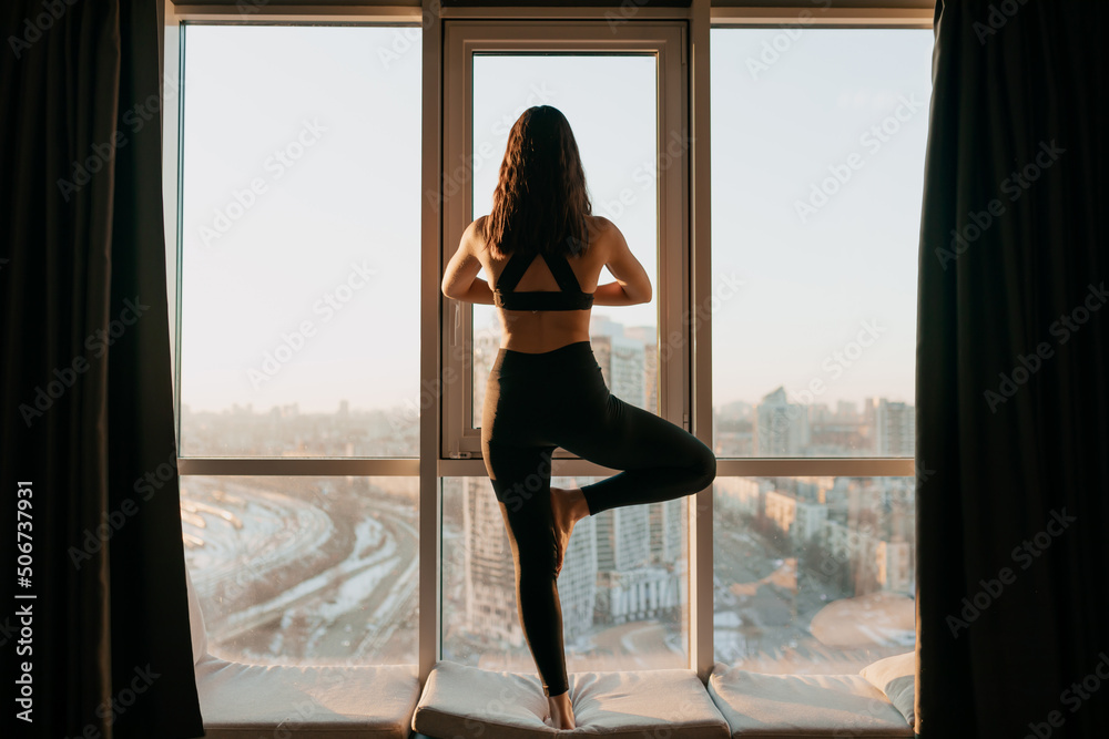 Full-lenght photo of attractive slim lady in uniform with long wavy hair doing yoga against window with city view in morning sunshine Full-lenght photo of attractive slim lady in uniform with long wavy hair doing yoga against window with city view in morning sunshine