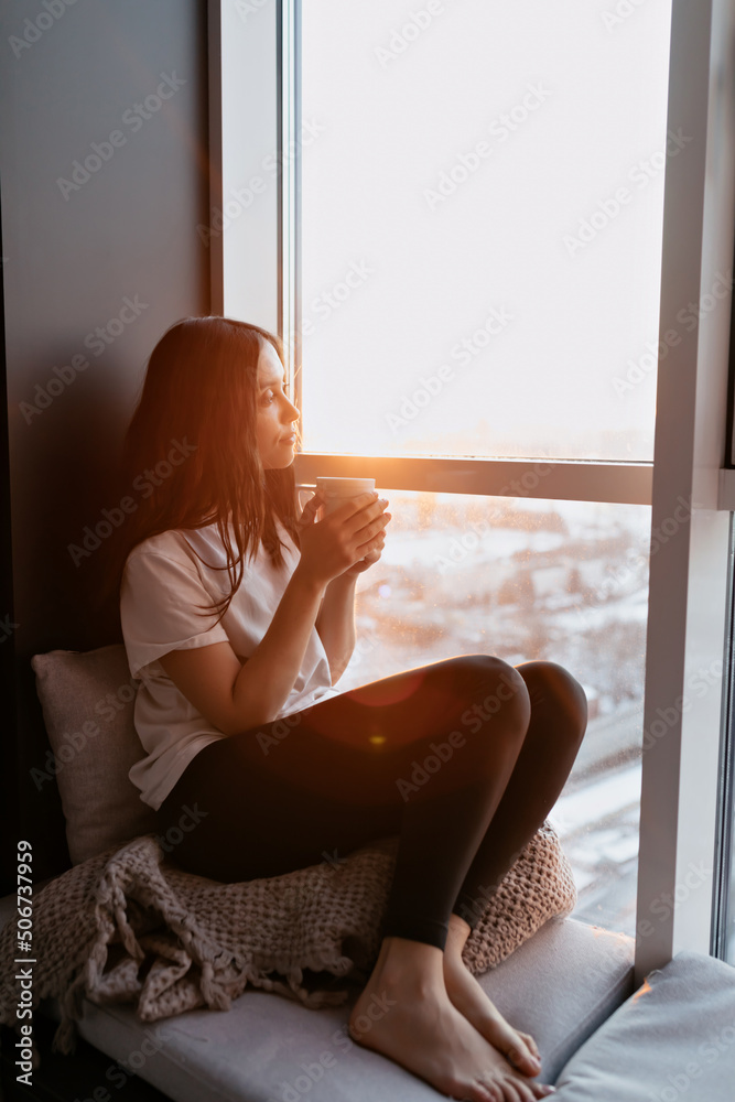 Full-length photo of relaxed charming lady with long wavy hair wearing white t-shirt and black leggings sitting near the window in sunlight on sunset with view on city