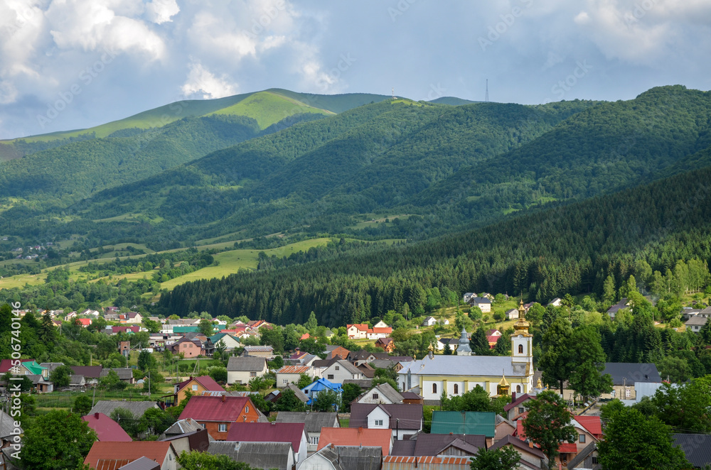 Beautiful rural landscape with colorful houses and church in the green meadow at the foot of the mountain ridge covered forest. Kolochava village in Carpathian Mountains, Transcarpathian, Ukraine