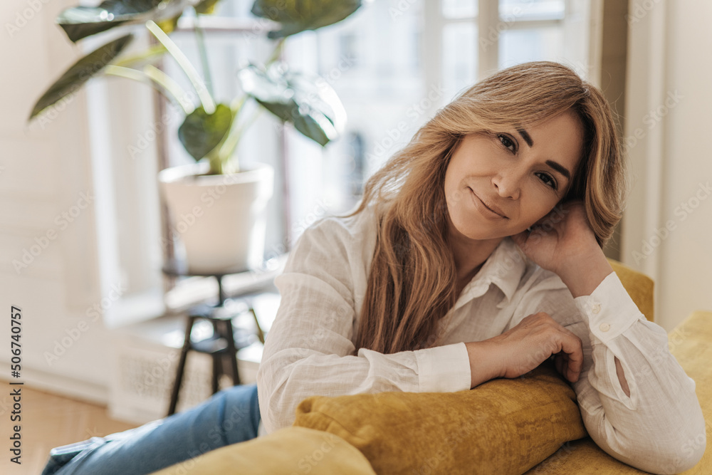 Beautiful middle-aged caucasian woman looking at camera while sitting on comfortable yellow sofa at home. Blonde wears white blouse while relaxing alone. Rest time concept. Beautiful middle-aged caucasian woman looking at camera while sitting on comfortable yellow sofa at home. Blonde wears white blouse while relaxing alone. Rest time concept.
