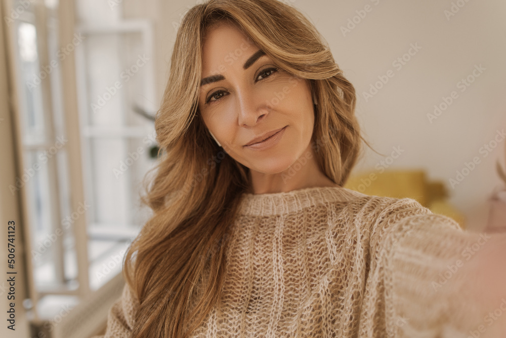 Pretty caucasian adult woman makes selfie imposingly looking at camera in light room. Blonde girl with long hair wears warm sweater. Lifestyle, female beauty concept Pretty caucasian adult woman makes selfie imposingly looking at camera in light room. Blonde girl with long hair wears warm sweater. Lifestyle, female beauty concept