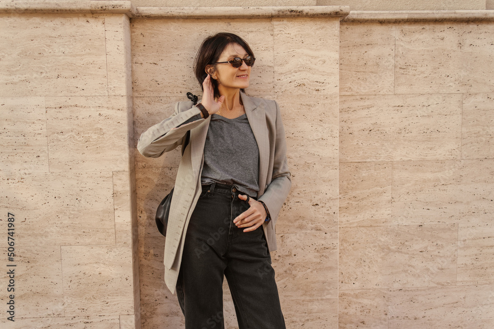 Caucasian adult cute woman with short dark haircut stands leaning against wall on street. Pretty woman in casual grey clothes and sunglasses. Weekend concept