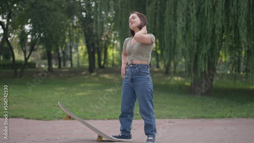 Wide shot confident Caucasian young woman with dwarfism standing with skateboard in spring summer park smiling looking away. Happy positive fit little person enjoying weekend leisure outdoors photo