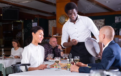 smiling male waiter serving order to adult man and woman in restaurante