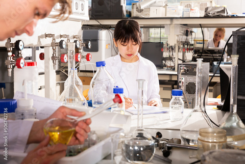 Young female Chinese chemist working in laboratory, writing report on results of chemical experiments in notebook