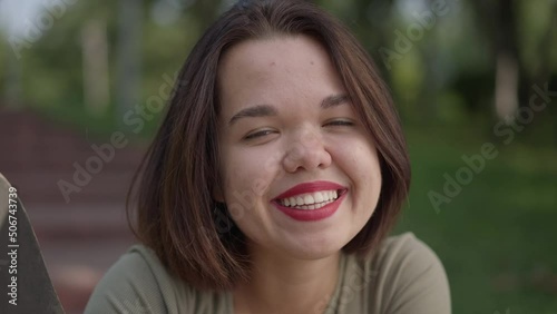 Front view close-up charming little woman smiling looking at camera sitting with skateboard in summer park. Portrait of happy confident fit Caucasian person with dwarfism posing outdoors photo