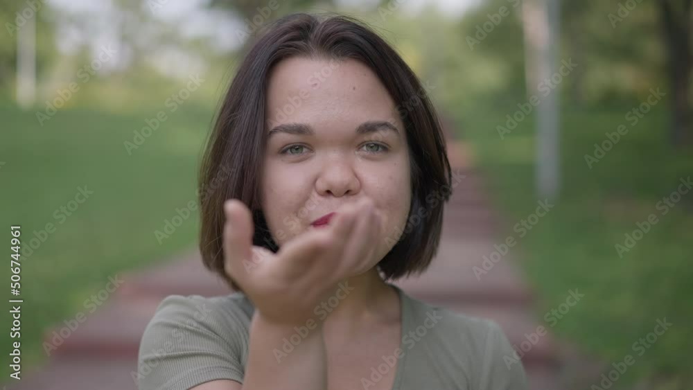Close-up coquette Caucasian little woman gesturing air kiss smiling looking at camera. Portrait of confident happy positive young person with dwarfism posing in slow motion flirting standing in park