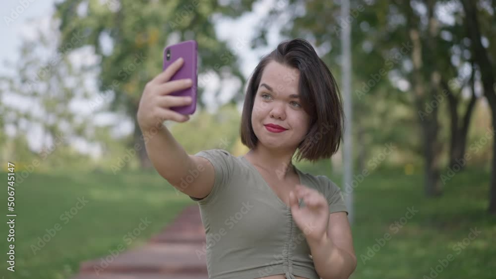 Beautiful little woman with dwarfism taking selfie on smartphone in slow motion outdoors. Portrait of smiling positive happy Caucasian little person photographing resting in park Beautiful little woman with dwarfism taking selfie on smartphone in slow motion outdoors. Portrait of smiling positive happy Caucasian little person photographing resting in park