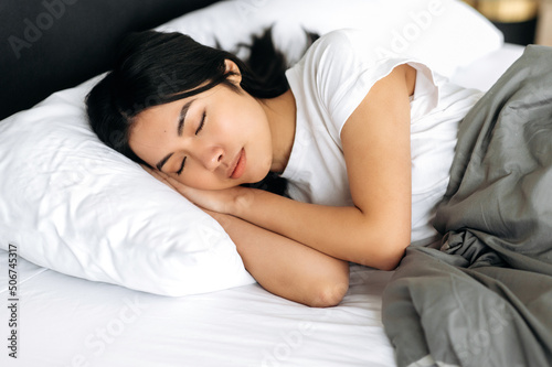 Side view of a sleeping asian girl in a cozy bed in apartment, pleasant dreams, healthy sleep. Pretty chinese woman in white t-shirt under the blanket in bed in the morning, sees sweet dreams