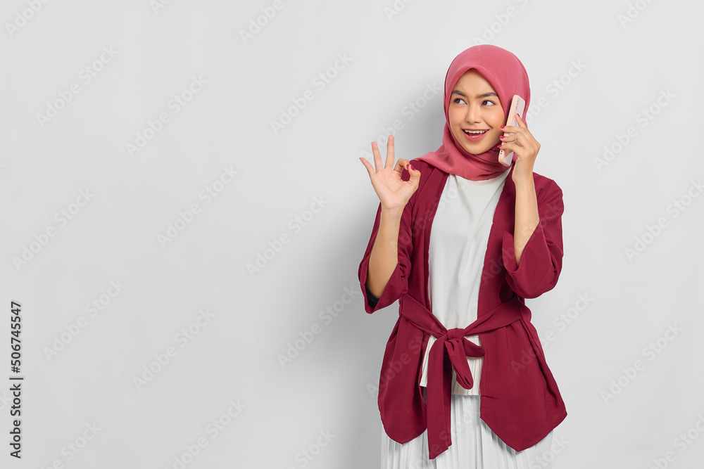 Cheerful beautiful Asian woman in casual shirt talking on mobile phone and looking away at copy space, showing okay sign gesture isolated over white background