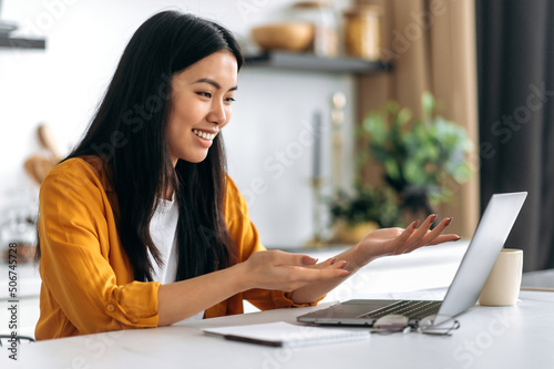 Friendly pretty asian brunette girl, manager, freelancer, sit in the kitchen, uses laptop, talking by video call with the client or employees, discussing project, holds consultation, smiling