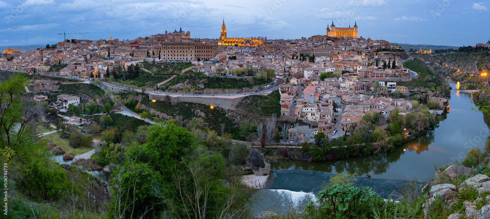 Picturesque twilight view of Toledo cityscape on green hilly banks of Tagus river in early spring overlooking illuminated ancient Cathedral with Gothic bell tower and fortified Alcazar castle, Spain..