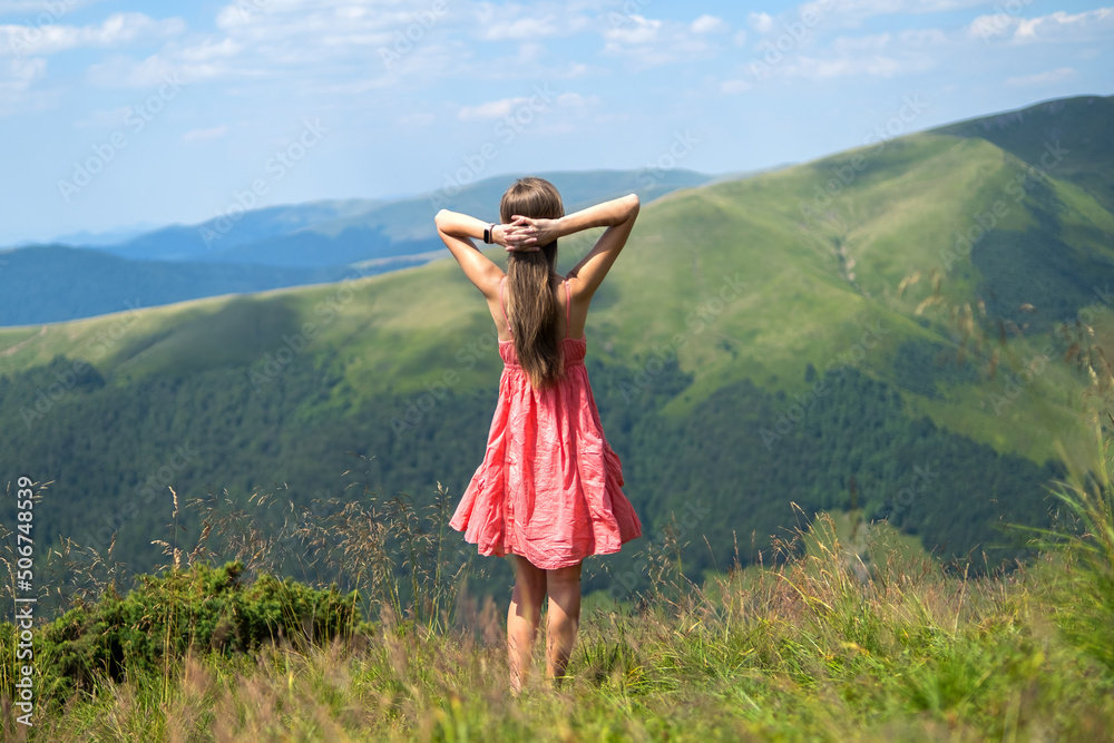 Young woman in red dress standing on grassy meadow on a windy day in summer mountains raising up her hands enjoying view of nature