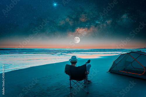 man sitting in a camping chair alone on the beach looking at the starry night next to a tent, back view