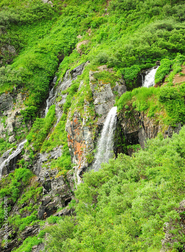 A small waterfall flowing down from a rocky mountain overgrown with dense shrubs.