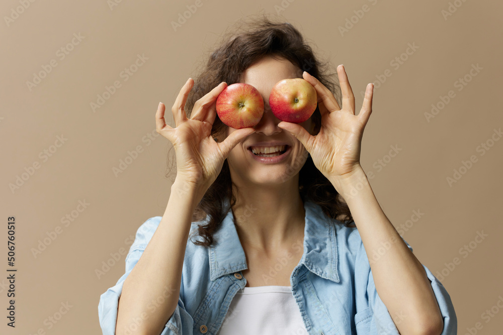 Funny cute curly female in jeans casual shirt closes eyes with apples smiles at camera posing isolated on over beige pastel background. Healthy food. Natural eco-friendly products concept. Copy space Funny cute curly female in jeans casual shirt closes eyes with apples smiles at camera posing isolated on over beige pastel background. Healthy food. Natural eco-friendly products concept. Copy space