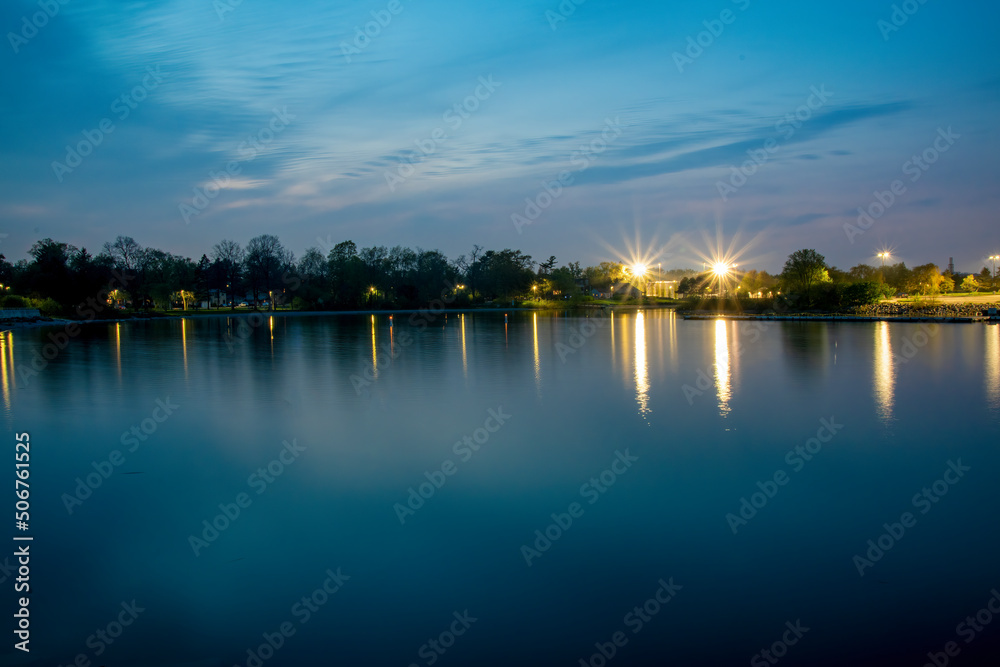RK McMillan Park in Mississauga, Ontario is seen across the glassy water of Lake Ontario at night time, taken with a long exposure.