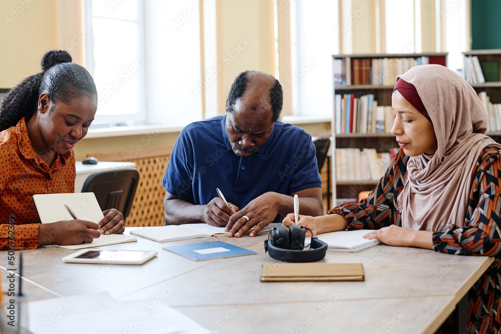 Group of three multi-ethnic immigrant students sitting at table in library doing writing task during lesson Group of three multi-ethnic immigrant students sitting at table in library doing writing task during lesson