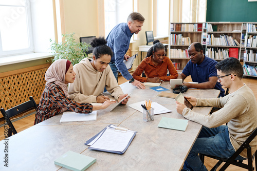 Mature Caucasian English language teacher helping immigrant students with doing task during lesson in library photo