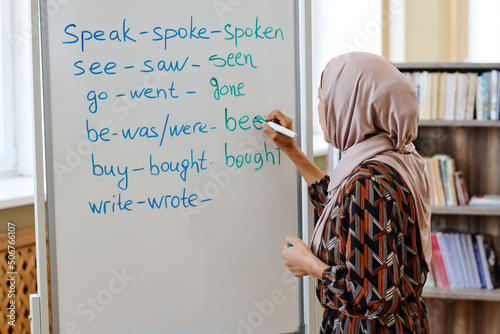 Unrecognizable woman wearing hijab standing at whiteboard doing task with irregular verbs during English language for immigrants lesson photo